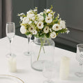 White flower bouquet in a glass vase on a dining table with candles and glasses.