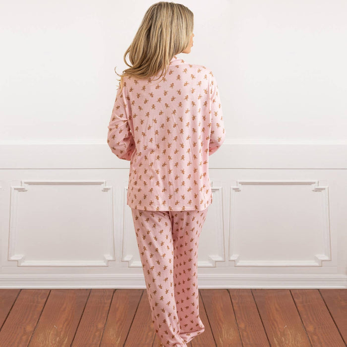 Woman wearing gingerbread patterned blush pink pajama set, standing indoors against white paneled wall