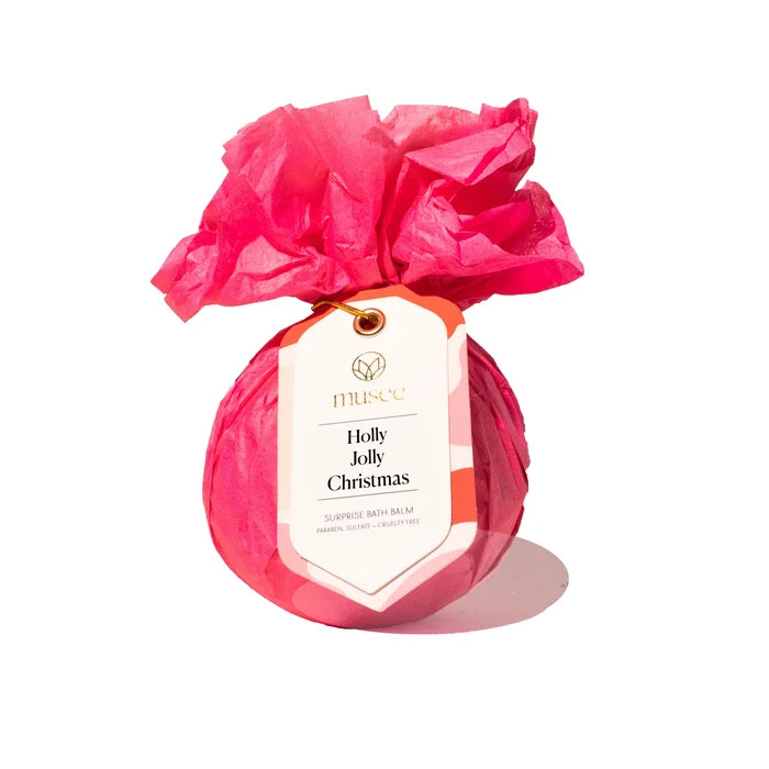 A round bath bomb wrapped in bright pink tissue paper and tied with a gold string, featuring a white paper tag that reads 'museé Holly Jolly Christmas Surprise Bath Balm' on a clean white background.