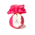 A round bath bomb wrapped in bright pink tissue paper and tied with a gold string, featuring a white paper tag that reads 'museé Holly Jolly Christmas Surprise Bath Balm' on a clean white background.