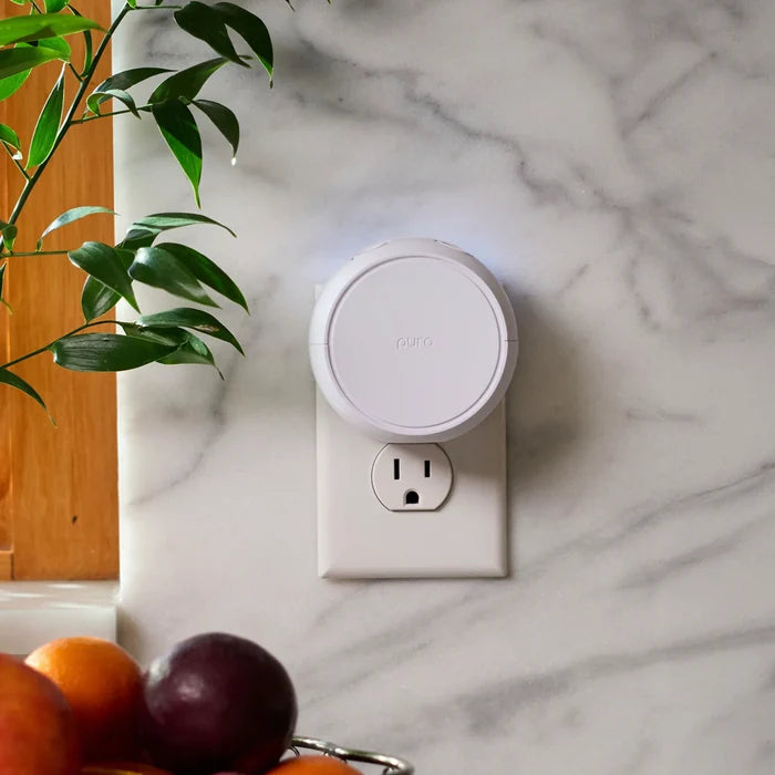 A white circular smart plug device with a glowing blue indicator light, plugged into a white wall outlet on a white marble surface. A wooden cabinet, green leaves, and fruit are visible in the background and foreground.