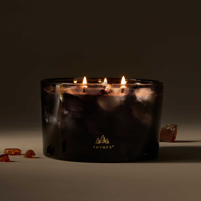 4-Wick 21 oz, A lit four-wick scented candle in a dark smoky glass jar with the THYMES logo printed in gold near the bottom. The candle is styled on a dark surface with a few amber-colored stones nearby against a dark background.